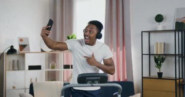 Evdeki koşu bandında çalışan ve cep telefonunda selfie çeken, kulaklıklı, yakışıklı, güler yüzlü, aktif, genç bir adamın portresi.