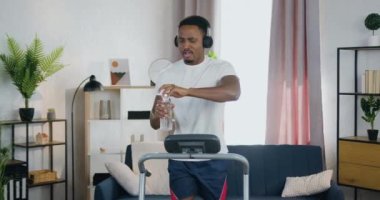 Koşu bandında koşan ve kulaklıkla müzik dinleyerek su içen yakışıklı, pozitif sporcu, siyah tenli bir adamın ev ödevi konsepti.