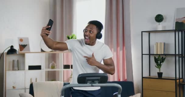 Портрет красивого улыбающегося современного активного молодого темнокожего парня в наушниках, который бегает по беговой дорожке дома и делает селфи на мобильном телефоне — стоковое видео