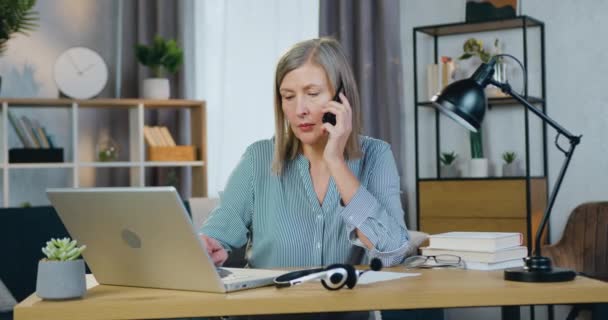 Старшая деловая женщина разговаривает по мобильному телефону и печатает на ноутбуке — стоковое видео