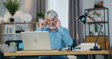 Gözlüklü emekli adam evde dizüstü bilgisayar kullanıyor.