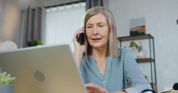 Mujer de edad que tiene charla móvil mientras trabaja en el ordenador portátil — Vídeo de stock