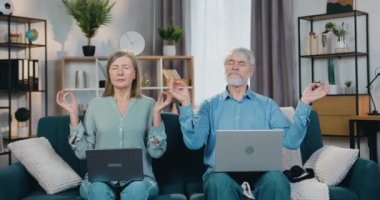 Laptoplarıyla kanepede meditasyon yapan barışçıl olgun bir çift.