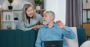 Kanepede dizüstü bilgisayarıyla oturan ve karısıyla konuşan yaşlı bir adam.