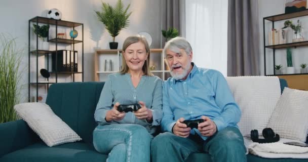 Возбужденная старшая пара, играющая в игры с беспроводными джойстиками — стоковое видео