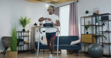 Ev işi konsepti, yakışıklı, sakin, kulaklıklı Afro-Amerikan genç bir adamın koşu bandında yürüdüğü ve şişeden su içtiği ve vücudundaki su dengesini tazelediği yer.