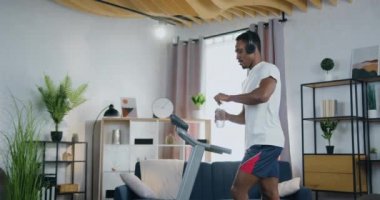 Keyfi yerinde, genç, siyah tenli, kulaklıkla müzik dinleyen ve evdeki koşu bandında antrenman yaparken su içen bir adam.