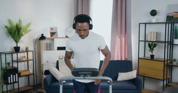 Guapo activo moderno casado de piel negra chico haciendo ejercicios de cardio en la cinta de correr en casa y escuchar canciones favoritas en los auriculares — Vídeo de stock