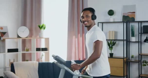 Cardio ασκήσεις στο σπίτι, όπου όμορφος χαμογελαστός ενεργός νεαρός μαύρο δέρμα τύπος με ακουστικά κοιτάζοντας κάμερα, ενώ το περπάτημα σε πίστα — Αρχείο Βίντεο
