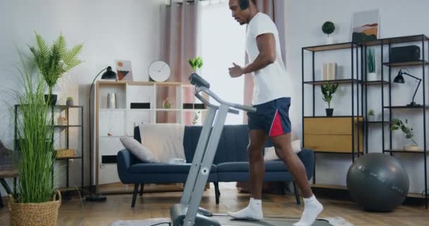 Vista lateral de boa aparência ativo moderno cara de pele preta em fones de ouvido que jogging na pista de corrida durante o treino em casa — Vídeo de Stock