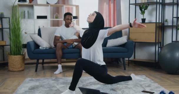 Aantrekkelijke ontspannen jonge vrouw in hoofddoek doen stretching oefeningen op de vloer in de buurt van haar zwart-gevilde man die hun zoon die probeert te slapen op vaders handen wiegen — Stockvideo