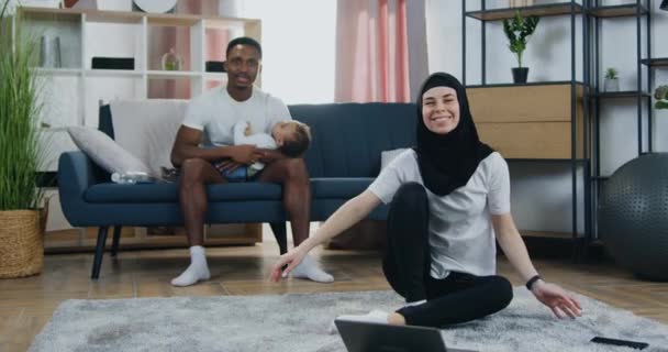 Pohledný usměvavý starostlivý černošky-kůže otec drží svého syna na rukou a dívá se do kamery se svou krásnou radostnou manželkou v hidžábu, který sedí na podlaze — Stock video