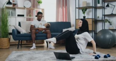 Çekici, sakin, genç, siyah tenli bir adam, küçük oğlunu sakinleştirirken, güzel, aktif, Arap karısı tesettürde, yerde esneme egzersizleri yapıyor.