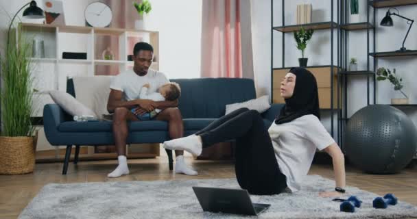 Aantrekkelijke kalme jonge man met een zwarte huid die zijn kleine zoon op handen neemt terwijl zijn vrij actieve Arabische vrouw in hijab oefeningen doet op de vloer — Stockvideo