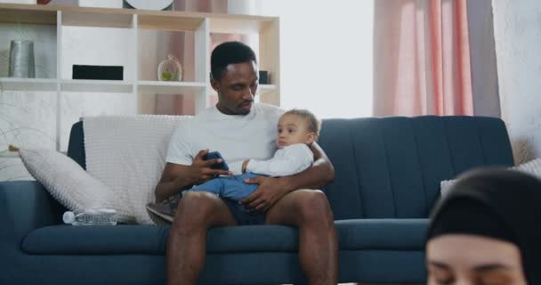 Guapo cariñoso padre afroamericano cariñoso mostrando dibujos animados en el teléfono inteligente para su hijo pequeño, mientras que su esposa activa bastante relajada en el hiyab haciendo ejercicios de estiramiento en el suelo — Vídeos de Stock