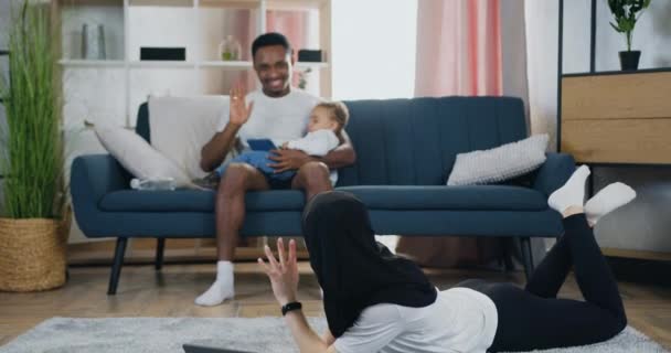Oldukça mutlu gülümseyen tesettürlü genç bir kadın dizüstü bilgisayarını kullanarak yerde yatıyor ve küçük çocuklarına bakan yakışıklı, pozitif, siyah tenli kocasına el sallıyor. — Stok video
