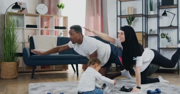 魅力的な積極的な現代の多様なカップルが自宅でストレッチ演習を行う健康的なライフスタイルの概念と、彼らの小さな息子がふわふわのカーペットの上で遊んでいる — ストック動画