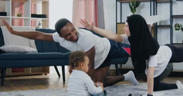 Adorable de bonne humeur actif aimant couple multiethnique faire des exercices d'étirement pendant l'entraînement à la maison et leur mignon petit fils à la peau foncée jouer près d'eux sur le sol — Video