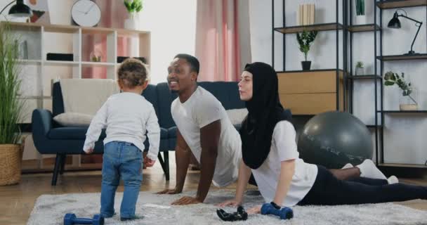 Guapo sonriente hombre de piel negra haciendo ejercicios deportivos junto con su esposa musulmana positiva en hijab en la alfombra cerca de su pequeño hijo en casa — Vídeos de Stock