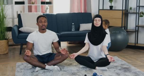 Attraktiv avslappnad modern skiftande par meditera med slutna ögon och korsade ben på golvet nära deras lekfulla lilla barn hemma — Stockvideo