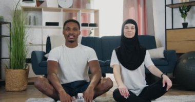 Çok güzel dekore edilmiş bir odada lotus poz veren ve kameraya başparmaklarını gösteren mutlu, evli, çok kültürlü çiftin ön görüntüsü.