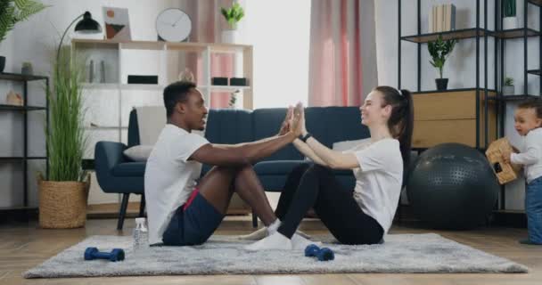 Концепция домашней тренировки, где привлекательные позитивные активные разнообразные пары делают упражнения для прессы с зажимами на полу, в то время как их темнокожий ребенок играет рядом — стоковое видео