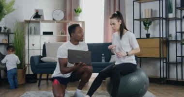 İyi görünümlü, genç, modern, sportif, karışık ırk çiftlerine yakın durun. Antrenmanda mola veriyorlar. Erkek, dizüstü bilgisayarda video eğitimi alırken kadın da spor salonunda su içiyor.