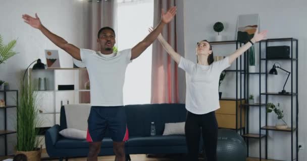 Aantrekkelijke positieve sportieve moderne diverse echtpaar doen ontspannende oefeningen en het beoefenen van yoga in prachtig ingerichte huis interier — Stockvideo