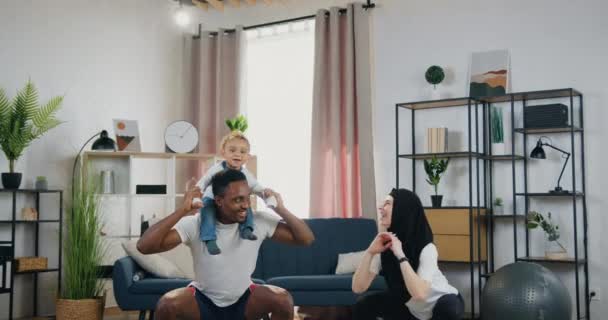 멋진 미소를 짓는 행복 한 스포츠 혼합 인종 커플 아버지 어깨에 어린 아이와 아늑 한 거실에서 웅크리고 운동하는 가정 운동 컨셉트 — 비디오