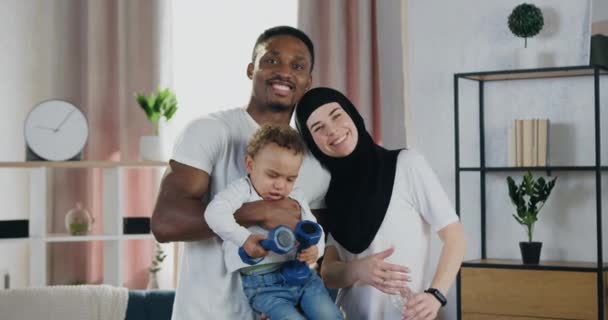 Aantrekkelijke glimlachende gelukkige multiraciale familie van Afrikaanse amerikaanse man en moslim vrouw en hun donkere huid kleine kind die op zoek naar camera thuis — Stockvideo