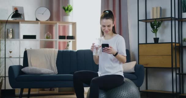 Portret van een mooie vrolijke glimlachende sportieve jonge vrouw in sportkleding die op fitnessbal zit, water drinkt en haar mobiel thuis gebruikt — Stockvideo