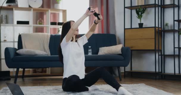 Attraktive aktive positive junge Brünette in Sportbekleidung sitzt zu Hause auf dem Boden und macht Sportübungen mit Stretching-Gummiband — Stockvideo