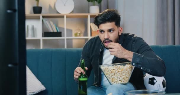 Отдых, спортивная концепция, где симпатичный возбужденный молодой бородатый парень внимательно смотрит спортивную игру по телевизору дома и ест попкорн, замедленная съемка — стоковое видео