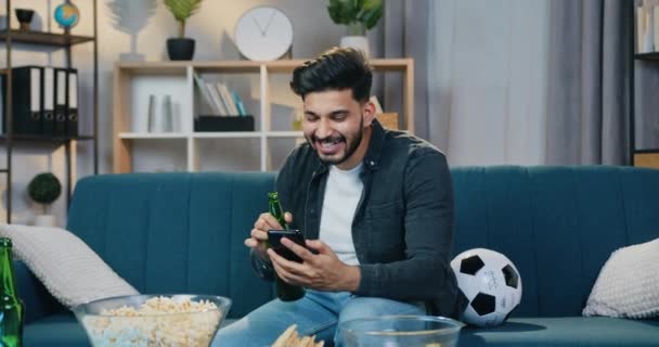 Agradable sonriente joven barbudo moderno sentado en el sofá en casa y hablando en el móvil a través de chat de vídeo con un amigo durante la visualización de partidos de fútbol en la televisión — Vídeos de Stock