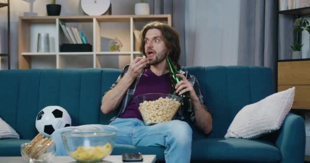 Aantrekkelijke positieve jonge mannelijke voetbalfan eten popcorn met bier en het maken van grote ogen bij het herzien van voetbalwedstrijd op tv thuis wanneer spelers een kans om doelpunt te scoren — Stockvideo