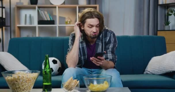 Handsome dissatisfied young bearded guy is sad while watching sport game on TV at home and starting to use his mobile to read news from friends — Stock Video