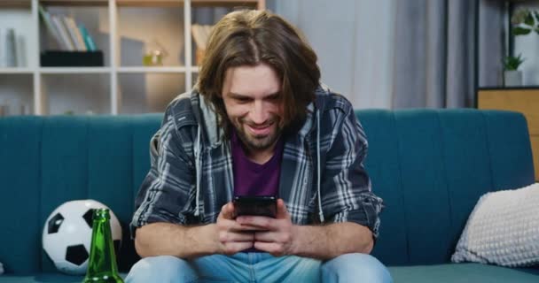 Vista frontal de un chico barbudo sonriente y tranquilo que se sienta en el sofá en casa y usa su teléfono inteligente para jugar, concepto de ocio — Vídeo de stock