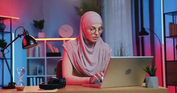 히잡에 있는 매력적 인 자신 감넘치는 실력있는 아라비안 호스 여성 이 집에 있는 직장에 앉아 다채 로운 야행성 효과를 보여 주는 노트북을 만들고 있다. — 비디오