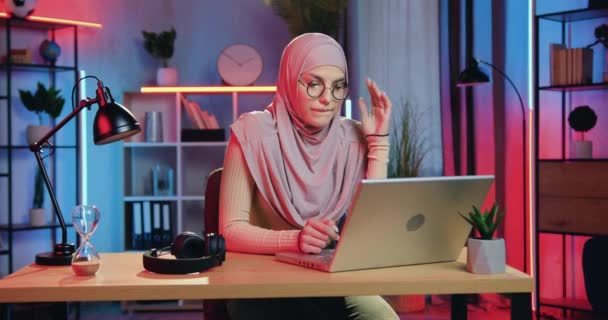 Atractiva mujer musulmana joven y segura de sí misma en hijab sentado frente a la computadora en la oficina en casa y trabajando por la noche, vista frontal, cámara lenta — Vídeos de Stock