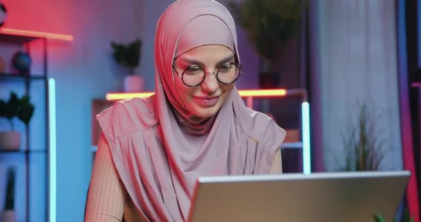 Közeli kép csinos mosolygós boldog fiatal muszlim lány hidzsáb szemüvegben, amely ül számítógép közelében színes megvilágított szoba és csevegés online barátaival — Stock videók