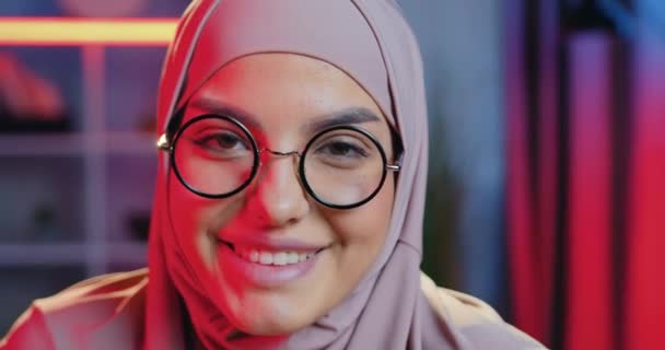 Primer plano de bastante feliz sonriente mujer árabe de 30 años en pañuelo para la cabeza que llevaba gafas y posando en la cámara con la expresión de la cara satisfecha en el fondo del interier habitación de la noche — Vídeo de stock