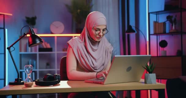 Vonzó pozitív képzett fiatal arab nő fejkendőben dolgozik laptop otthon este, és nézi a kamera őszintén mosolyog — Stock videók