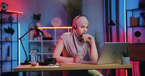 Piękny pewny siebie nowoczesny muzułmanin kobieta w hidżabie nosi słuchawki e-learning podczas słuchania online webinar w domu w godzinach wieczornych — Wideo stockowe