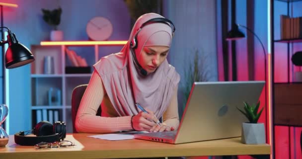 Vonzó komoly magabiztos 25 éves arab nő fejkendővel visel fejhallgatót az online webinar során, és fontos megjegyzéseket tesz a notebookba — Stock videók