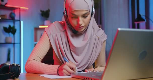 Espléndida mujer arábiga inteligente atenta y confiada en hijab lleva auriculares inalámbricos escuchando la presentación en línea en el ordenador en casa por la noche y escribiendo notas importantes — Vídeos de Stock