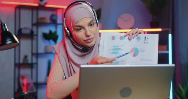 Adorable positivo seguro hábil musulmana joven en hijab explicando gráficos a través de video reunión en el ordenador portátil sentado en la oficina en casa por la noche — Vídeos de Stock