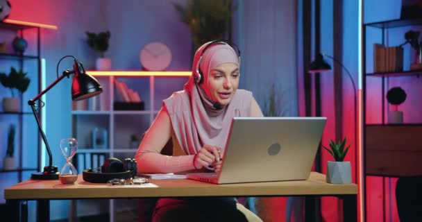 Belle jeune femme musulmane amicale et souriante en hijab donnant des conseils à son collègue lors d'un chat vidéo depuis son bureau le soir, concept de travail à distance — Video