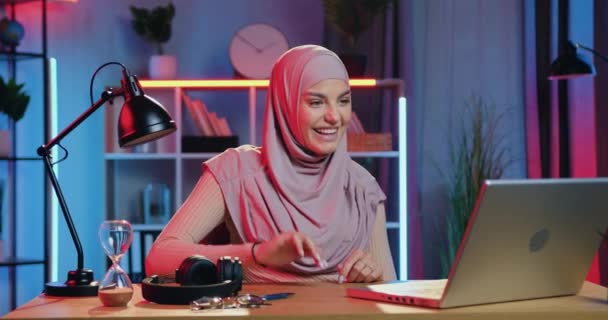 Retrato de la encantadora alegre chica árabe de 30 años feliz en pañuelo para la cabeza que se sienta frente a la computadora en la acogedora habitación iluminada por la noche y hablando por videollamada con un amigo — Vídeos de Stock
