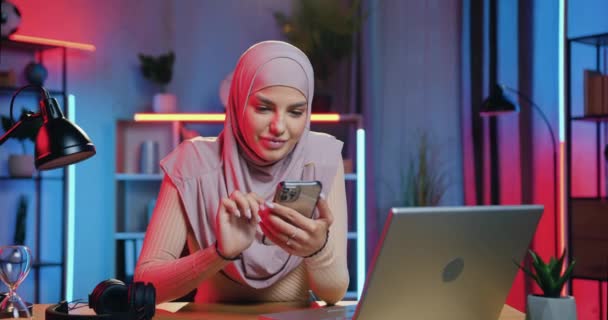 Mooie glimlachende gelukkig moderne jonge moslim vrouw in hijab maakt gebruik van haar smartphone tijdens het maken van pauze in haar werk op laptop in huis kantoor in de avond — Stockvideo