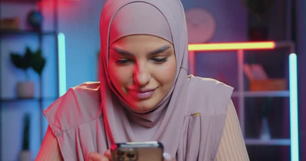 Zasněný krásný pozitivní mladý muslim žena v hidžábu sedí v barevné osvětlené místnosti ve večerních hodinách a pomocí smartphone psaní sms na své přátele — Stock video